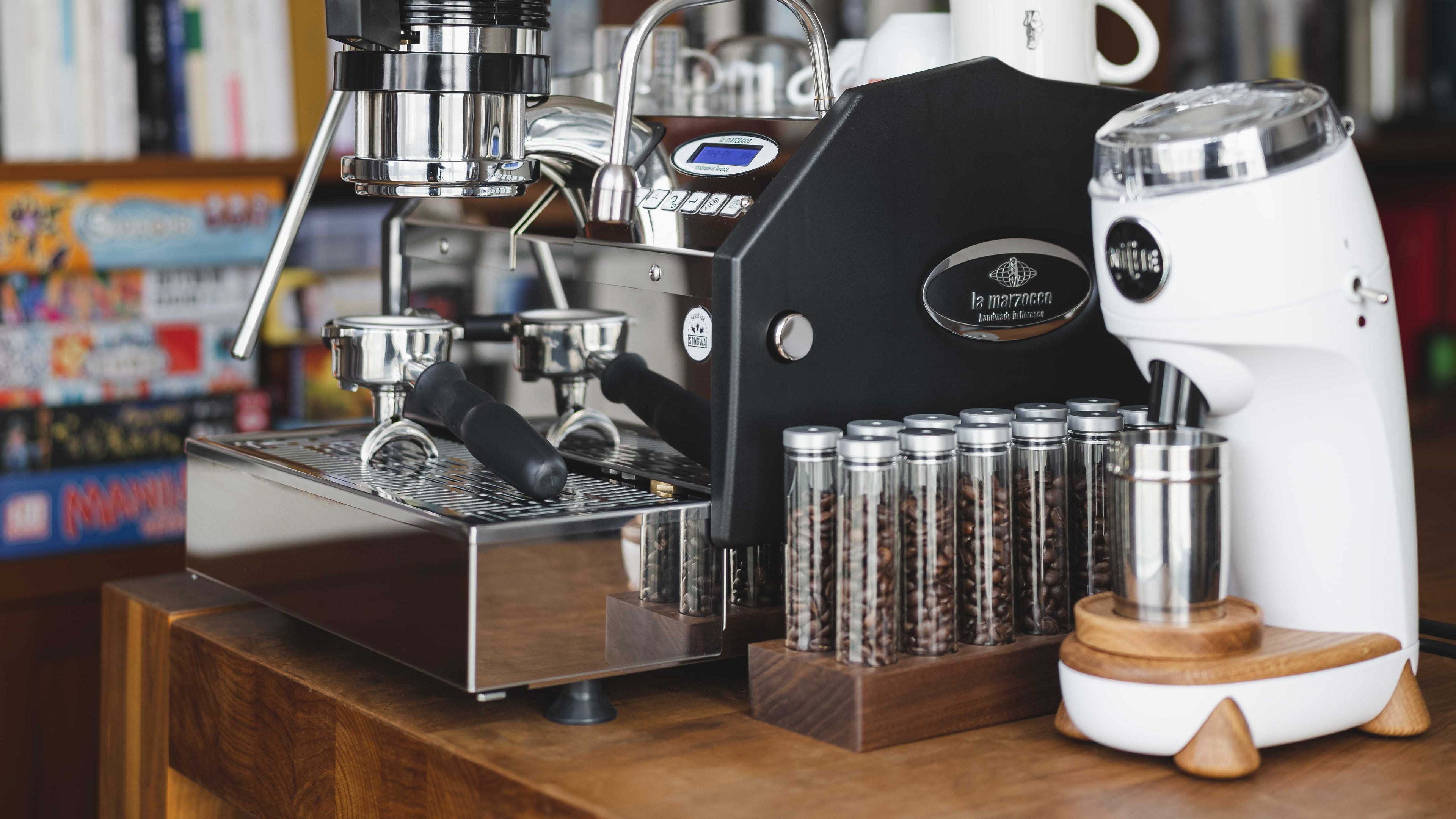 A high-end coffee station setup featuring a black and stainless steel espresso machine, a white coffee grinder, and several glass canisters of coffee beans, arranged on a wooden counter in front of a bookshelf.